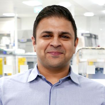 A man in a light blue shirt smiling in a bright office setting.