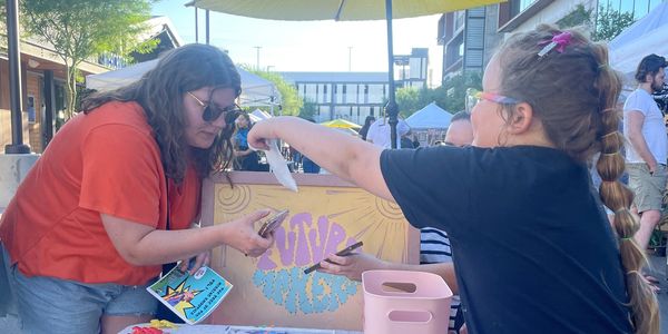 A girl hands something to a woman at an outdoor market stall.