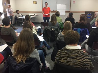 A woman standing in front of a crowd seated with their backs to the camera. SFPD podium nearby.