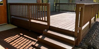 A wooden deck and stairs attached to a house with green siding and a brown door.