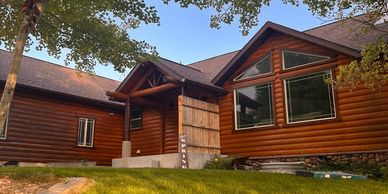A cozy log cabin surrounded by green grass and leafy trees under a clear sky.