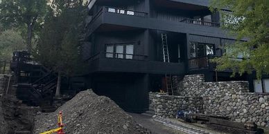 Modern black house under construction with stone walls and a dirt mound in front.