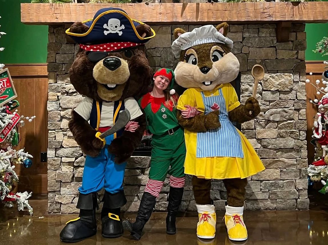Two animal mascots and a person in a festive elf costume pose happily indoors.
