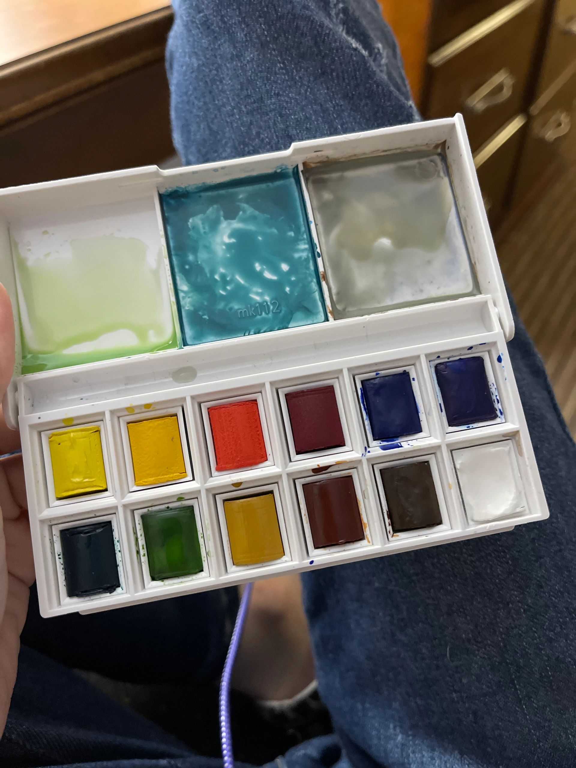 A small watercolor paint palette with various colors held over denim jeans.