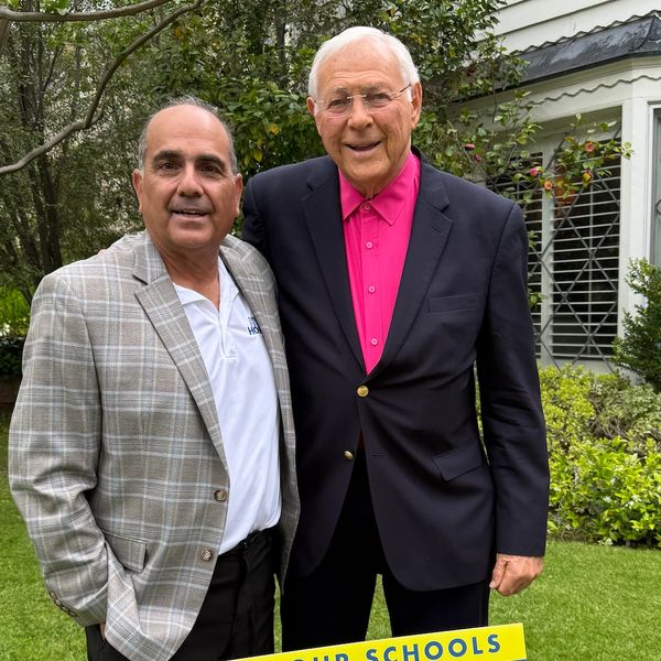 Two men smiling outdoors in front of greenery and a house.