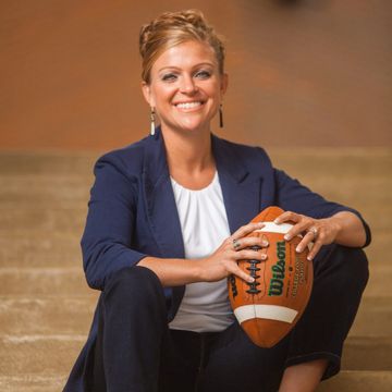A smiling woman in a blazer & white top sits on the floor with a football against a warm backdrop.