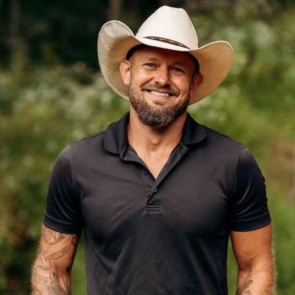 Man outdoors wearing a black polo and cowboy hat, smiling with hands in pockets.