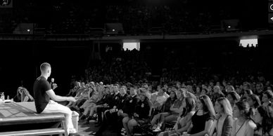 Large audience watching a speaker on a brightly lit stage.