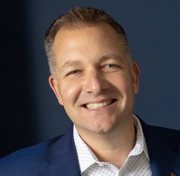 Man in a blue jacket smiling against a dark blue backdrop.