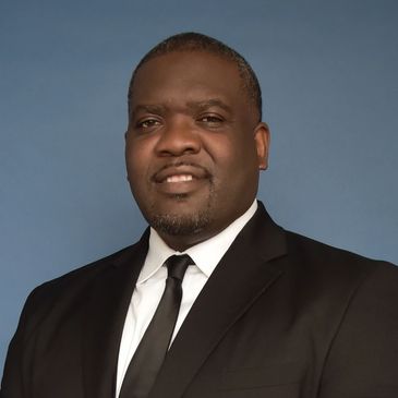 A confident man in a black suit and tie against a blue background.