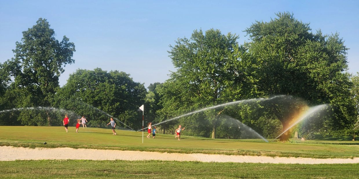 Children playing on a golf course with sprinklers and a rainbow.