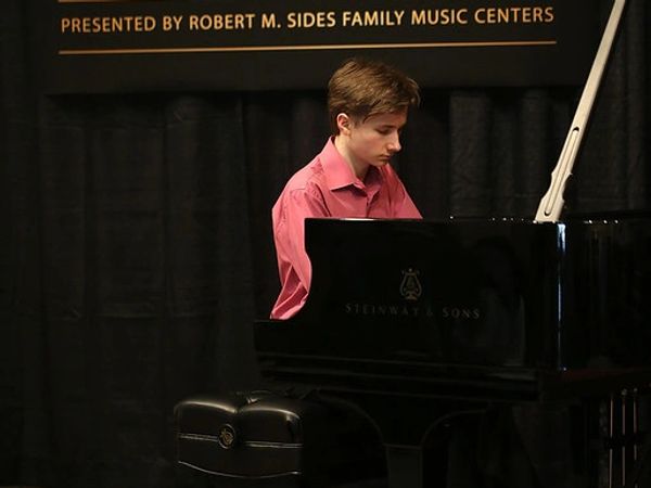 A photo of Michael in a pink shirt facing away from the camera while playing a black grand piano.