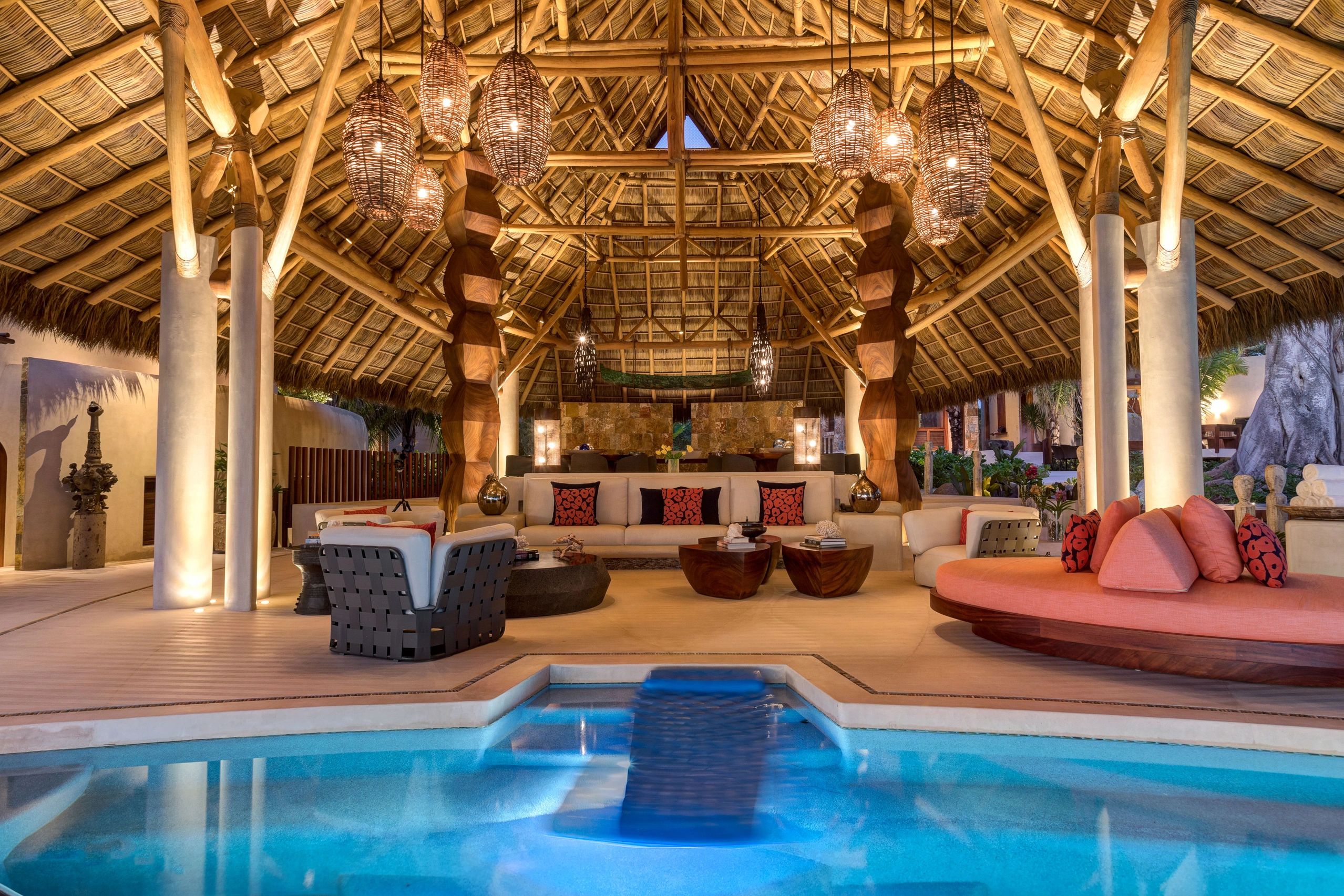 Palapa iluminada con luz cálida, resaltando techos y muebles, complementada por la piscina brillante.