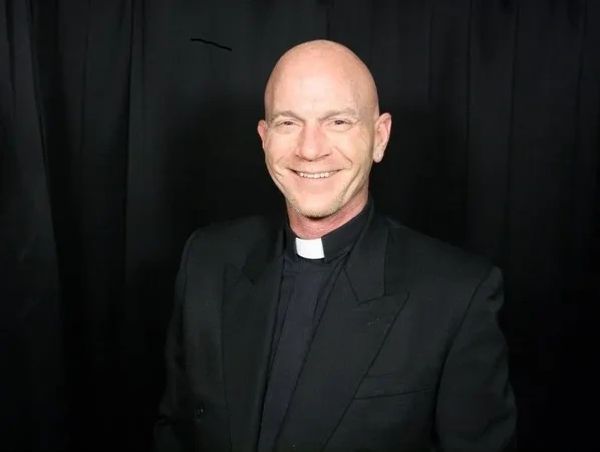 Smiling bald man in a black clerical suit with a white collar on a black background.