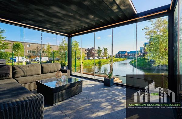 Modern outdoor seating area with glass walls overlooking a canal and greenery.