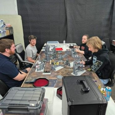 Four men playing a tabletop strategy game with miniatures and terrain.