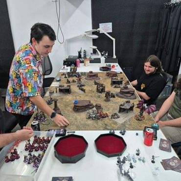 Three people playing a tabletop miniature game with terrain and figures.
