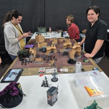 Three people engaged in a tabletop miniature war game with detailed terrain and figures.