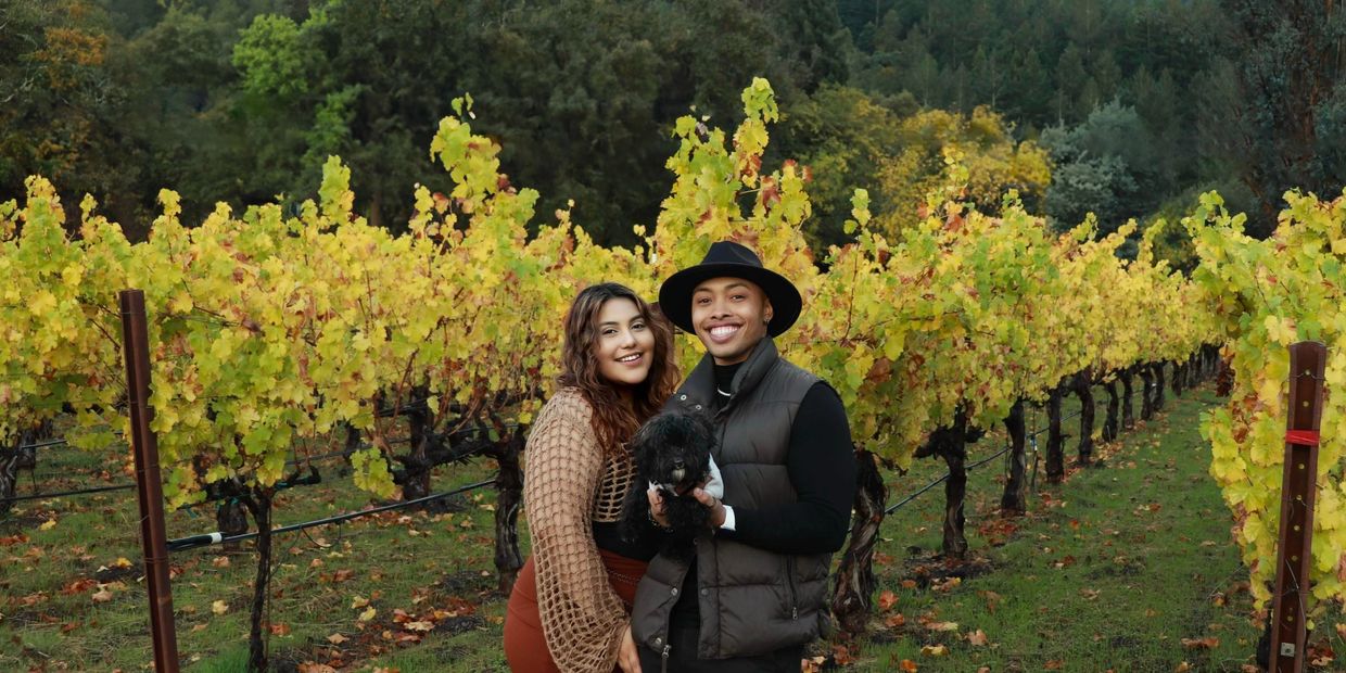 Couple smiling with their dog in a vineyard.