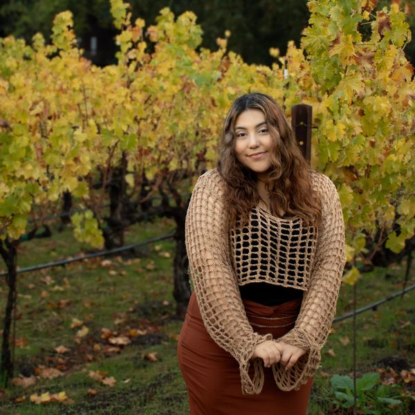 Woman smiling in a vineyard wearing a brown crochet sweater.