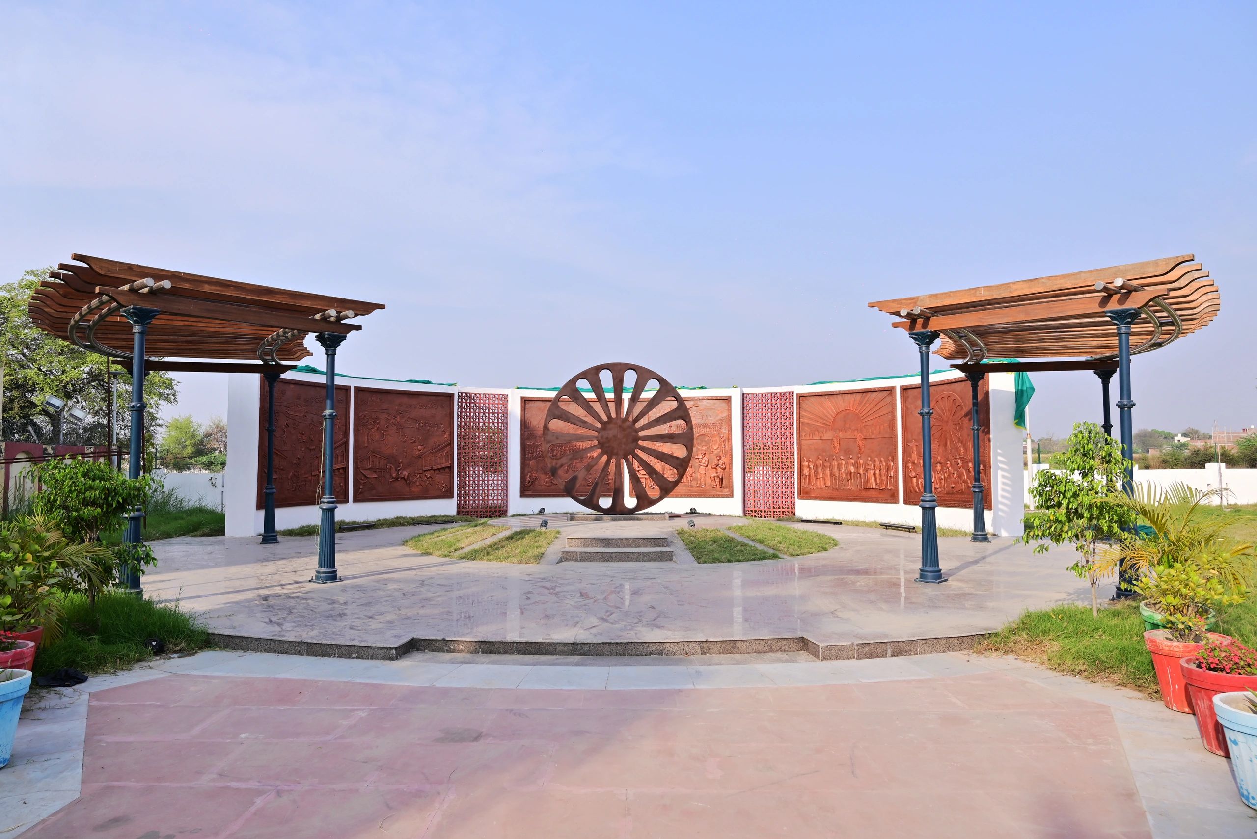 Open-air memorial with a large wheel sculpture and wooden pergolas.