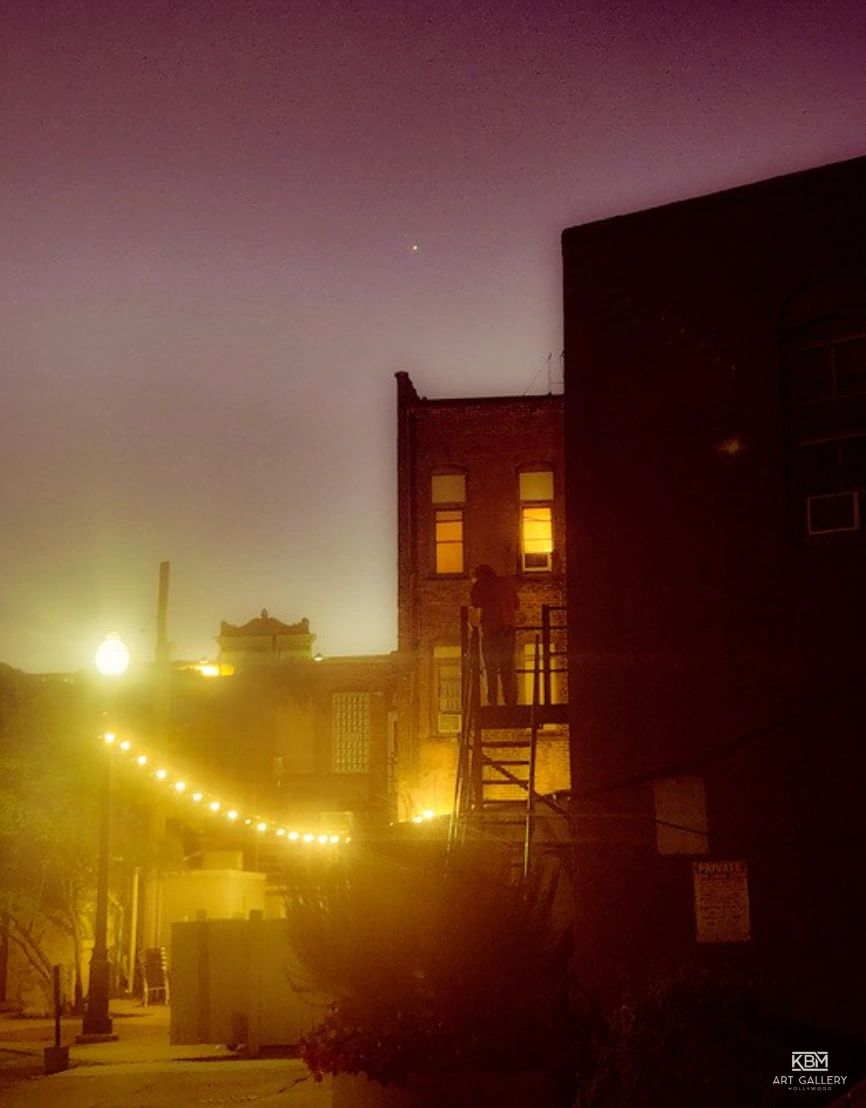 A dimly lit urban alley with string lights and a person standing on a fire escape at dusk.