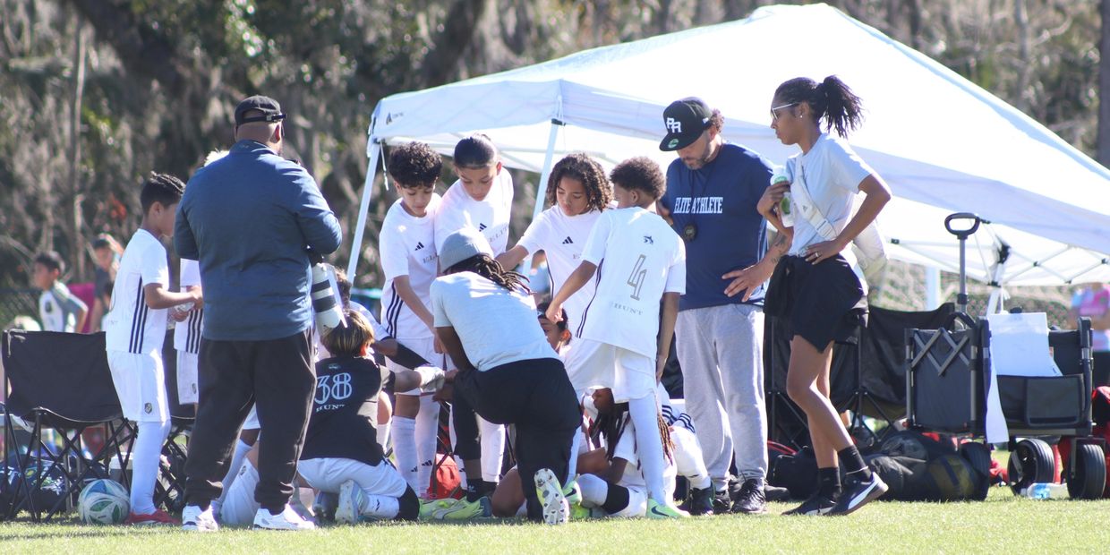 Youth soccer team gathers around coaches during a game break under a canopy.