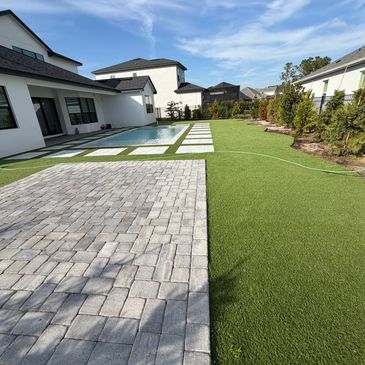 Modern backyard with pool, artificial grass, and stone patio under a blue sky.