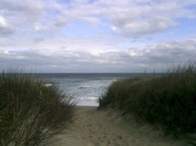 Pathway through sand dunes to the ocean under a cloudy sky.
