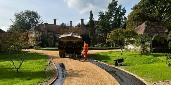 Worker preparing a gravel driveway with a truck and wheelbarrows on a sunny day.