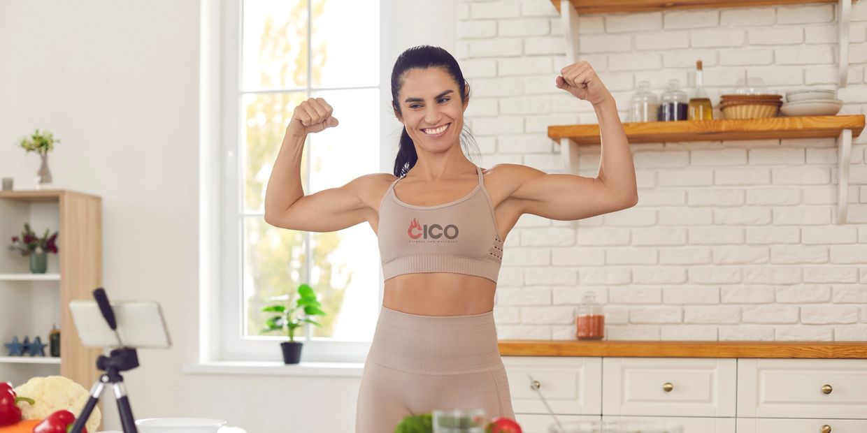 Strong woman flexing in kitchen wearing CICO Fitness top, promoting healthy lifestyle and nutrition coaching