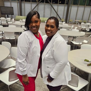 Two smiling women in white coats posing in a modern room with round tables and chairs.