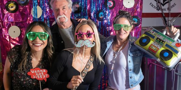 Four adults in fun props posing at a vibrant party photo booth.