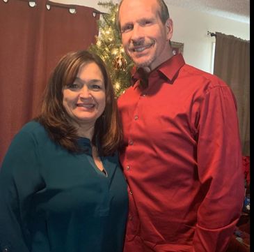 A smiling couple stands together in front of a decorated Christmas tree indoors.
