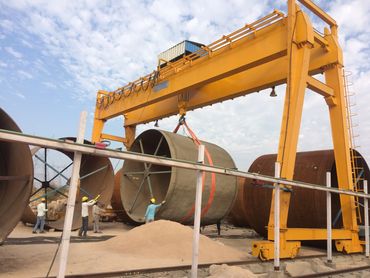Workers use a yellow gantry crane to lift a large cylindrical concrete structure outdoors.