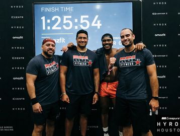 Four men in fitness shirts pose proudly in front of a finish time board.