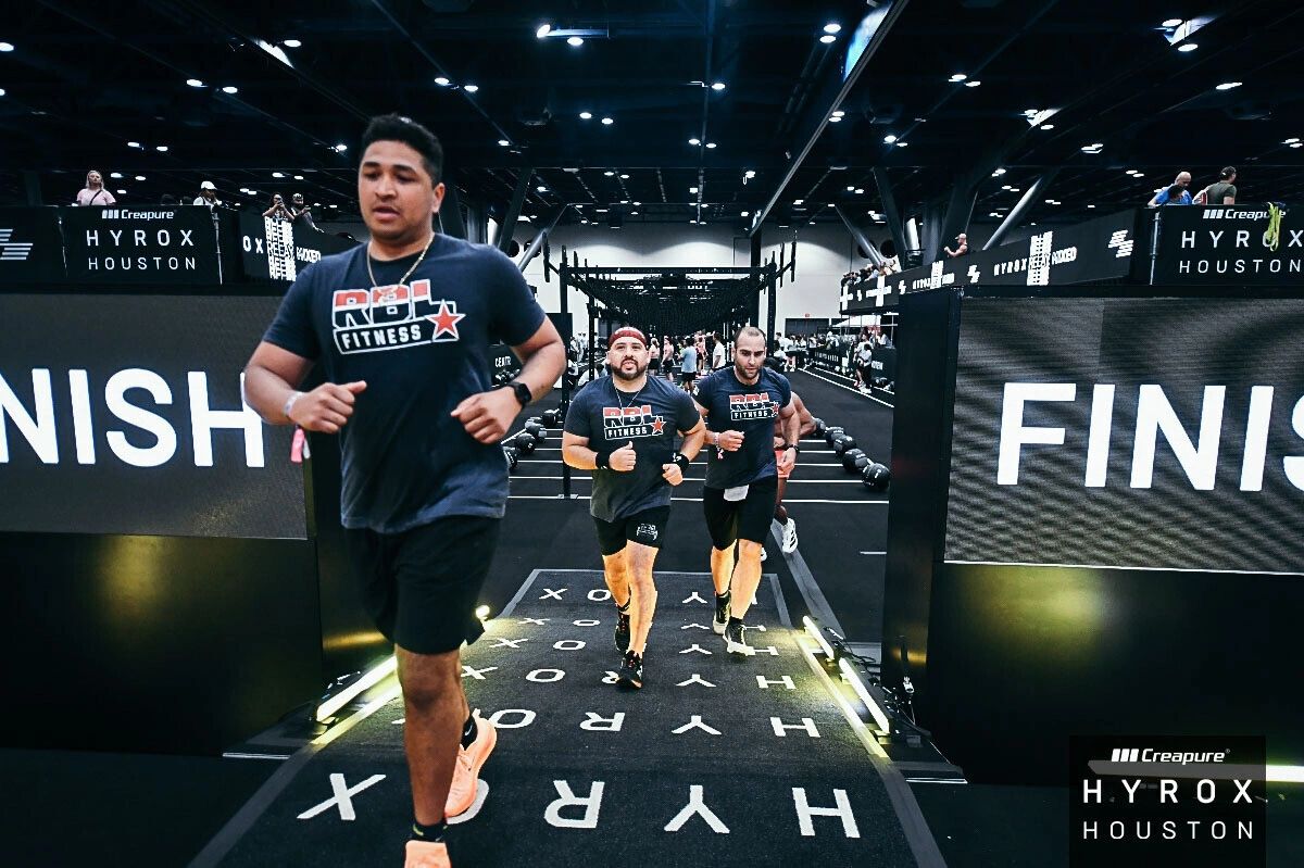 Three men running towards the finish line at a HYROX fitness event in Houston.