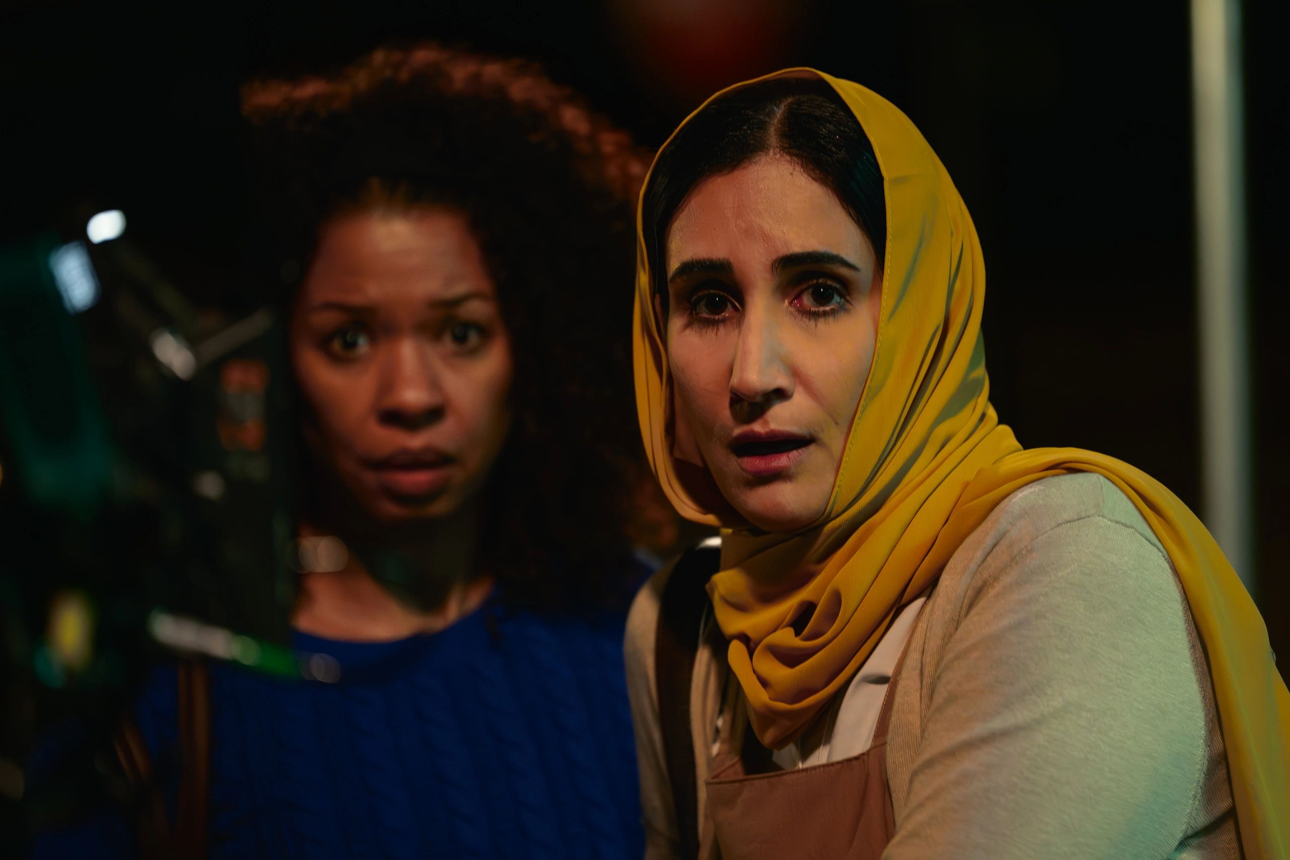 Two women, one in a yellow headscarf, look worriedly at something off-camera in dim lighting.