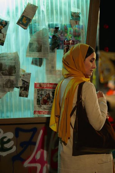 Woman in yellow headscarf stands pensively by a window covered with hanging newspapers.