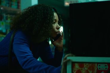Woman in blue shirt talks on phone in dimly lit room.