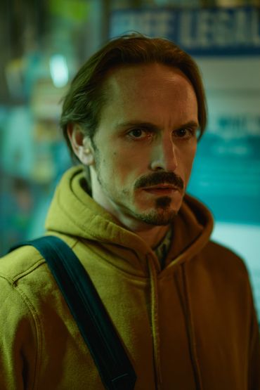 Man with intense expression wearing a yellow hoodie and shoulder strap.