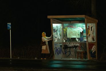A person waits at a graffiti-covered bus stop at night.