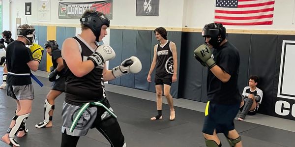 Two people sparring in a martial arts gym with protective gear and others training nearby.