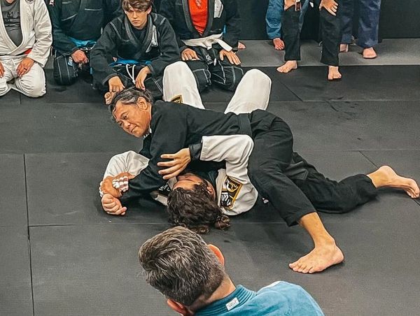 Two people practicing Brazilian jiu-jitsu on mats with onlookers.