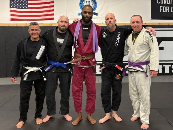 Five men in Brazilian Jiu-Jitsu gis posing on a mat with an American flag in the background.