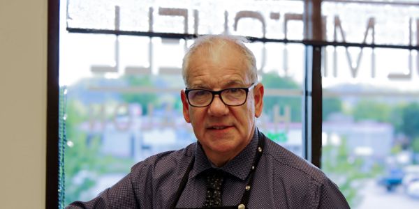 An elderly man with glasses smiles indoors near a window.