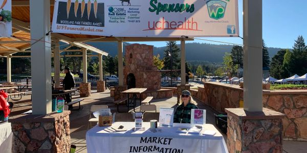 Market information booth at a sunny outdoor farmers market with a person sitting behind the table.
