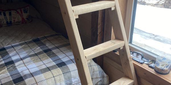 Wooden ladder leaning against a bed with a plaid blanket near a window.