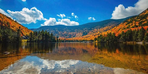 Clear lake reflecting autumn trees and blue sky with clouds in a mountain landscape.