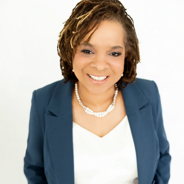 Confident woman in a navy suit smiling warmly with pearl necklace and hands in pockets.